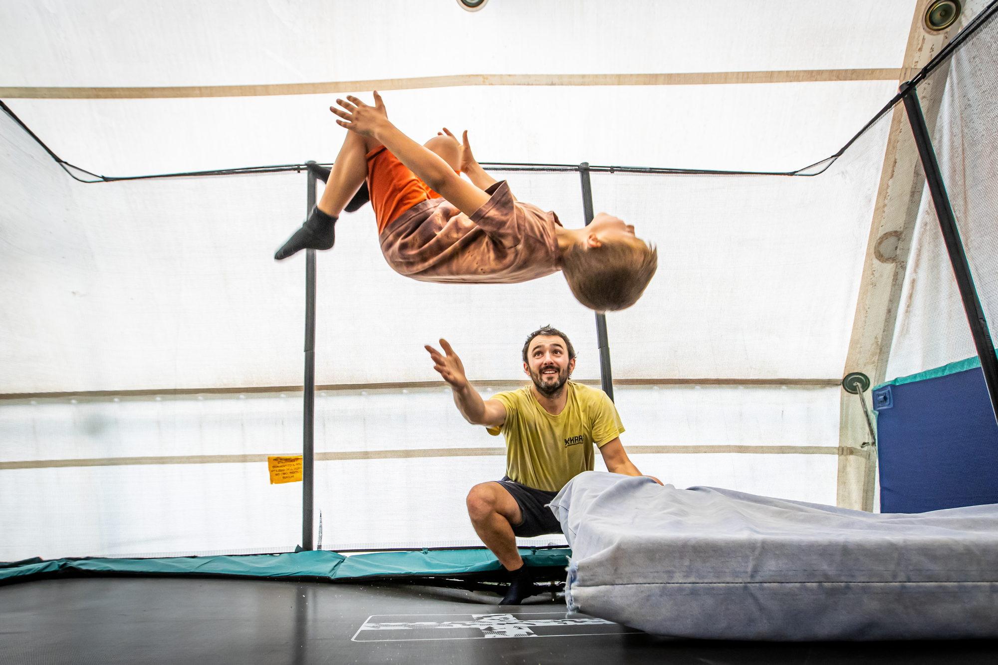 Un bambino a mezz'aria in un salto all'indietro sul tappeto elastico, e l'istruttore che lo segue con le mani pronte